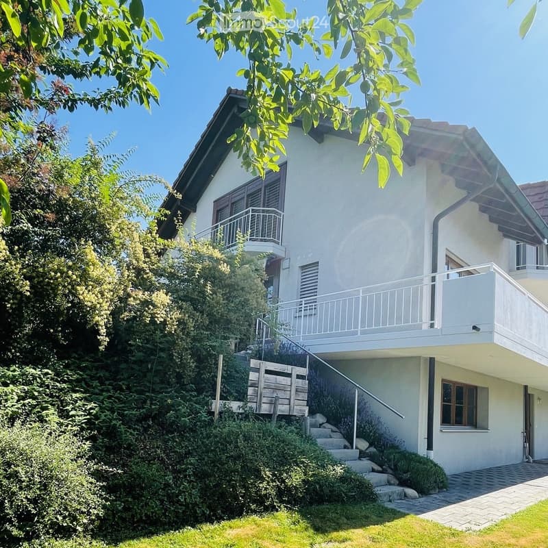 Two-story white house with balconies, surrounded by greenery and a well-maintained lawn.
