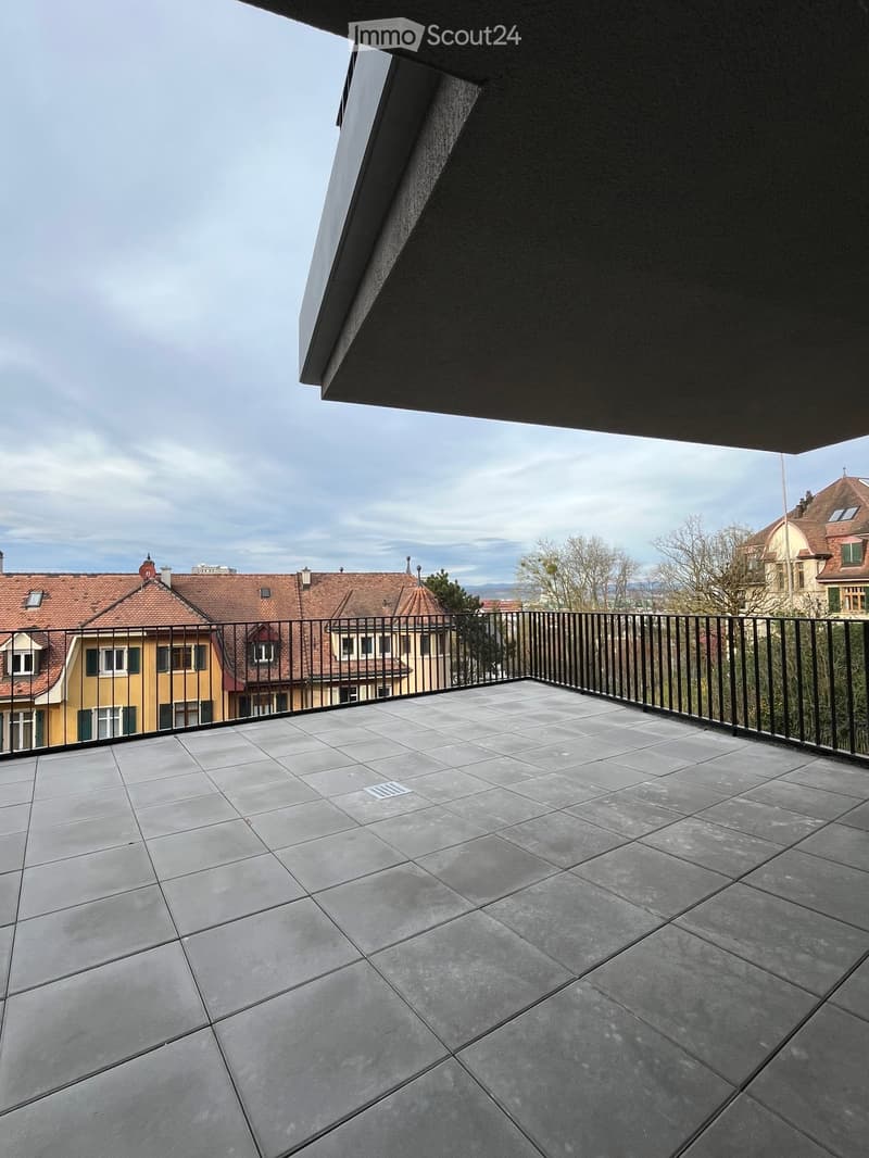 Balcony with grey tiled floor, black metal railing, overlooking cityscape with traditional buildings and trees.
