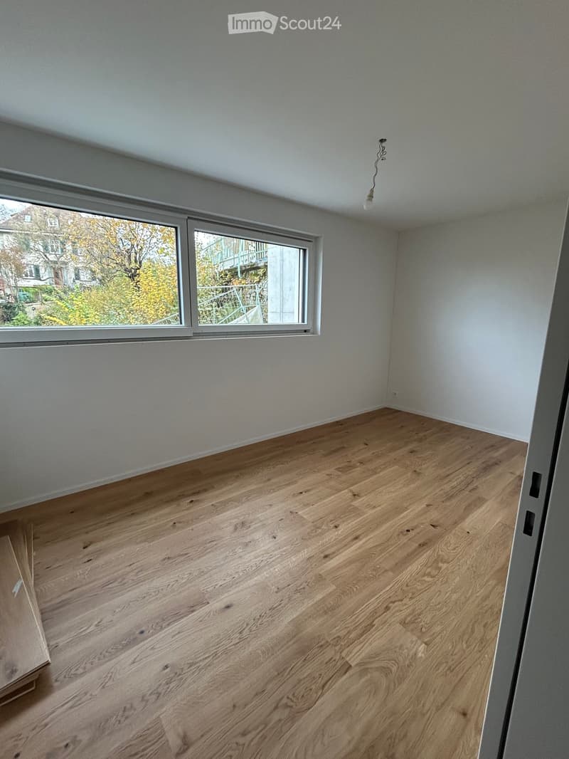 Empty room with light wooden flooring and white walls, featuring large windows with a view of the outside.