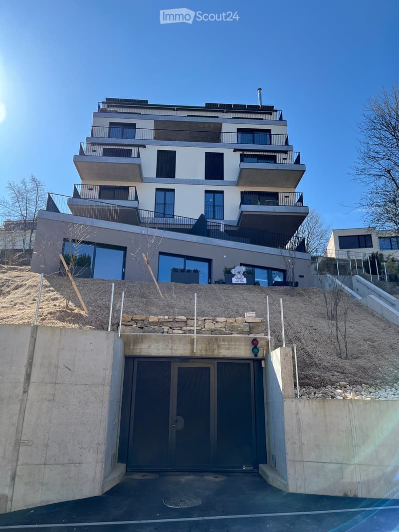 Modern multi-story building with balconies, large windows, and a raised ground floor leading to a garage entrance.