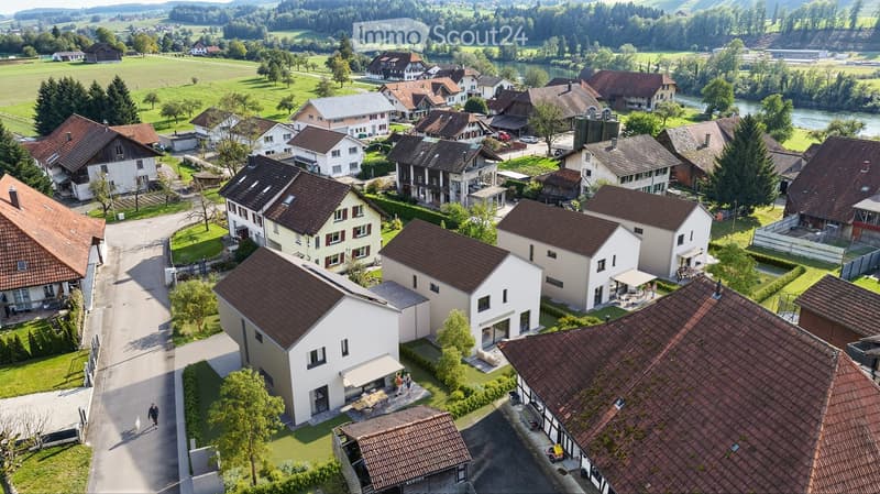 Aerial view of a residential area with multiple houses, surrounded by green fields and a river in the background.
