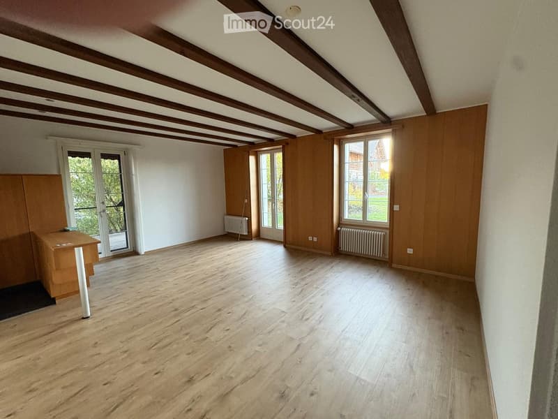 Empty room with wooden flooring, exposed wooden ceiling beams, large windows, and a sliding glass door leading outside.