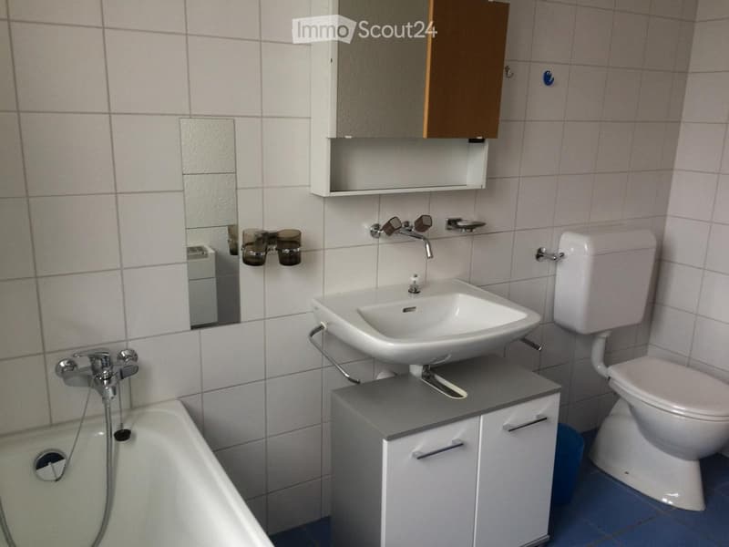 Modern bathroom with white tiles, a sink, toilet, bathtub, mirror, and a small cabinet.