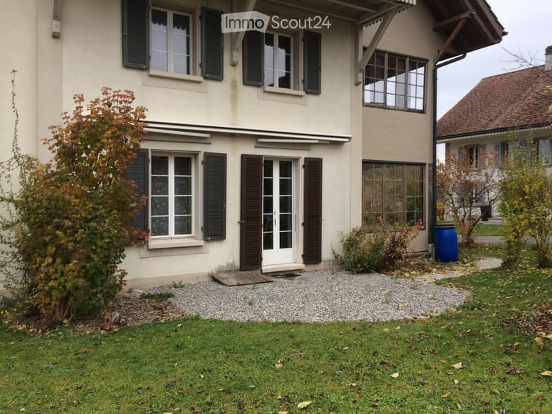 Two-story house with beige exterior, multiple windows with shutters, a front door with glass panels, and a small garden area with grass and gravel.