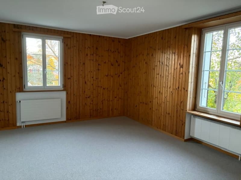 Empty room with wooden panel walls, carpeted floor, two windows, and radiators under the windows.