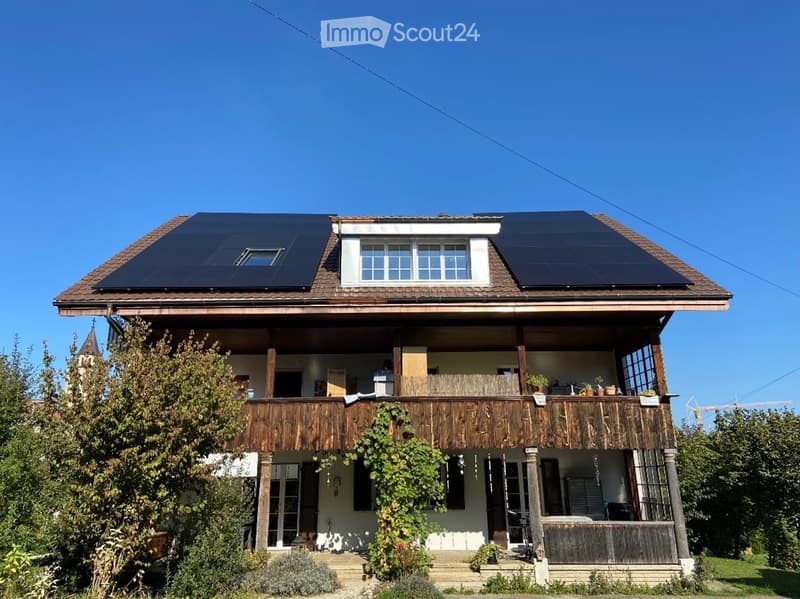 Two-story house with a wooden balcony, solar panels on the roof, and surrounding greenery.