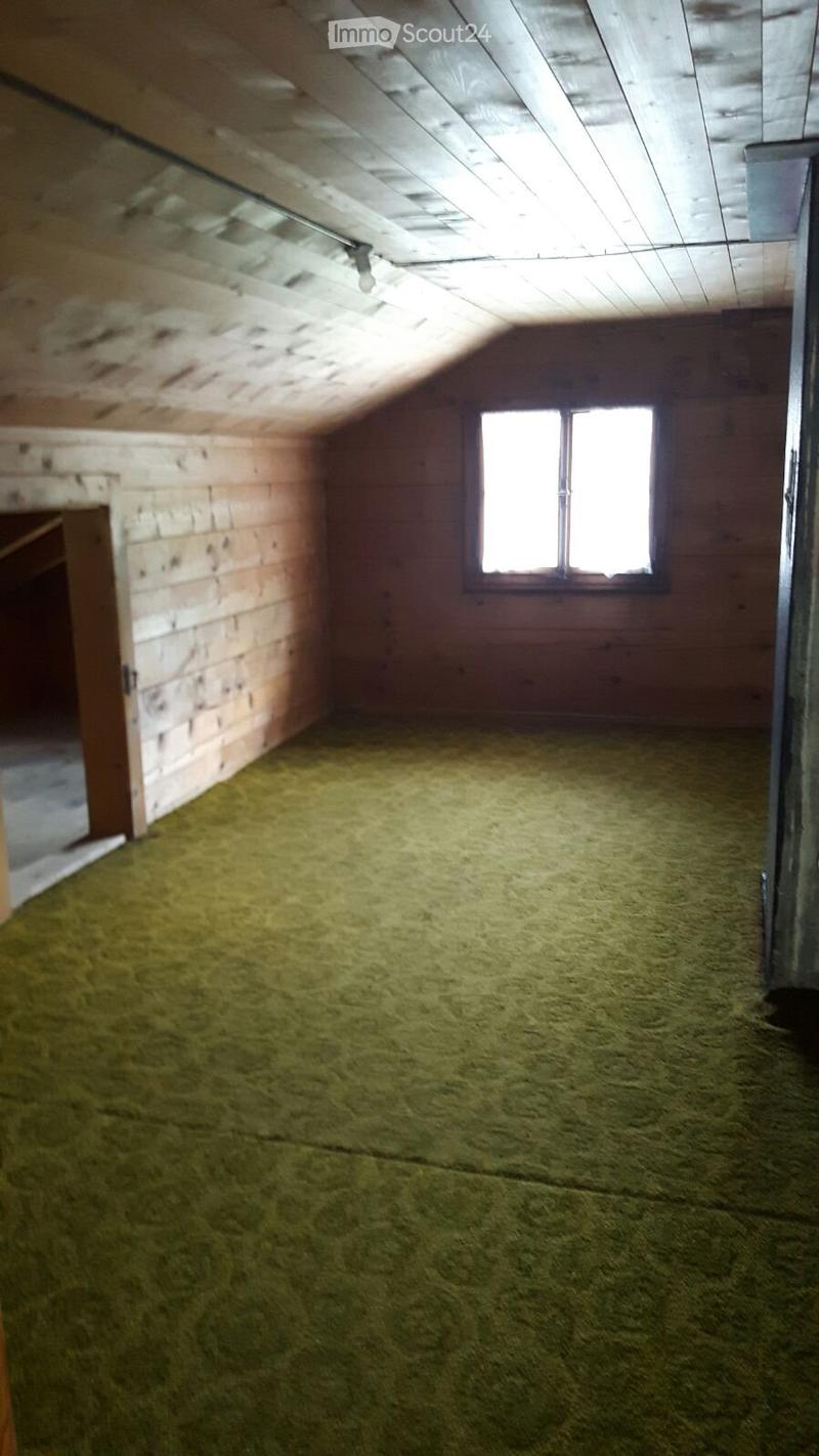 Empty attic space with wooden walls, exposed wooden ceiling beams, and a single window letting in natural light. The floor is covered with straw or hay-like material.