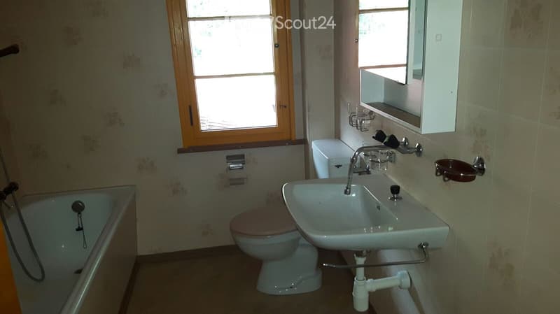 Bathroom with a bathtub, toilet, sink, mirror, and window with wooden frame. Light-colored tiles and floral wallpaper.