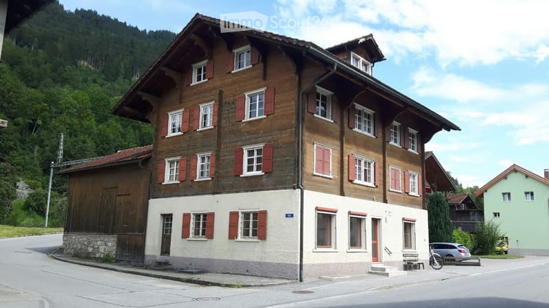 Traditional wooden house with red shutters, multiple windows, and a stone base, situated in a scenic, mountainous area.