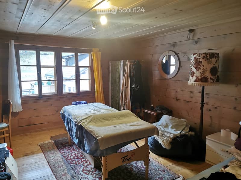 Wooden-paneled room with a massage table, chair, mirror, floor lamp, and small table. Large windows provide natural light.