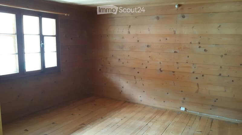 Completely empty room with wooden walls and floor, featuring a single window and a power outlet on the wall.
