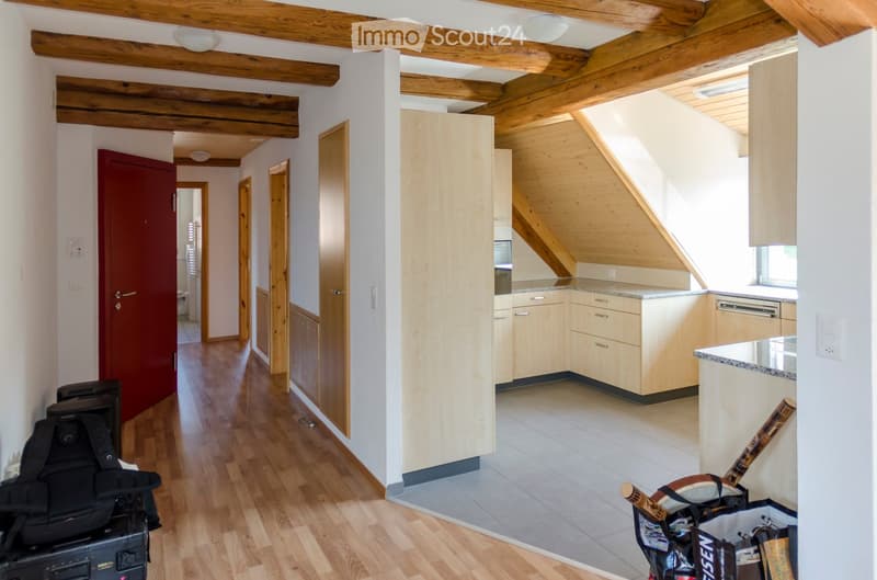 A modern, well-lit hallway leading to a kitchen area with light wooden cabinets and a granite countertop. The hallway features exposed wooden beams and hardwood flooring, with a backpack and suitcase placed near the entrance.