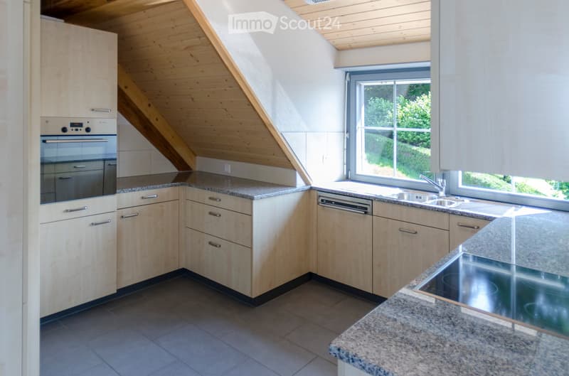 Modern kitchen with light wood cabinets, granite countertops, stainless steel appliances, and a large window overlooking greenery.