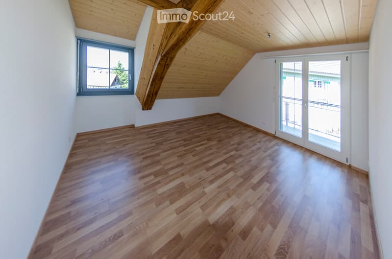 Empty attic room with wooden flooring, white walls, exposed wooden beams, and large windows and sliding doors leading to a balcony.