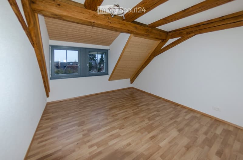Empty attic room with wooden floors, white walls, exposed wooden beams, and a single window offering an outside view.