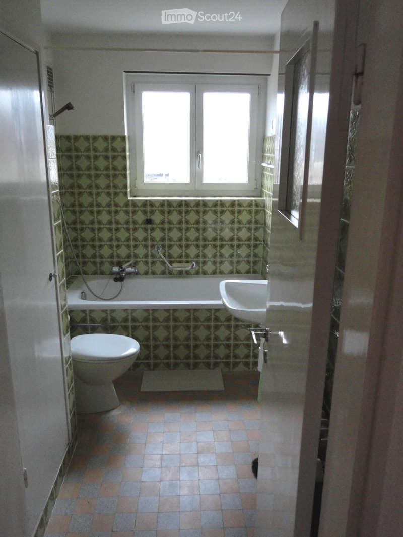 Bathroom with green patterned tiles, bathtub, toilet, sink, and a window above the bathtub.