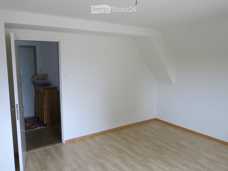 Empty room with light wooden flooring and white walls, featuring an open doorway leading to another room with a small cabinet and some folded items.