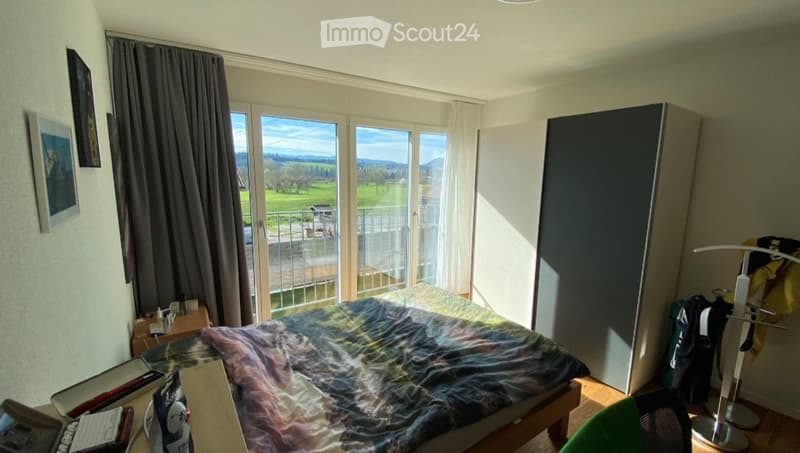 Bright bedroom with large windows offering a scenic view, featuring a bed with a colorful blanket, a nightstand, a wardrobe, a clothing rack, and a chair.