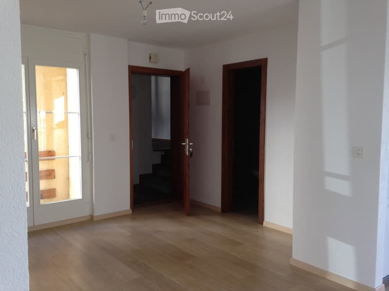 Empty room with wooden floor, white walls, two doors, one with a glass window, and a staircase visible through the open door.