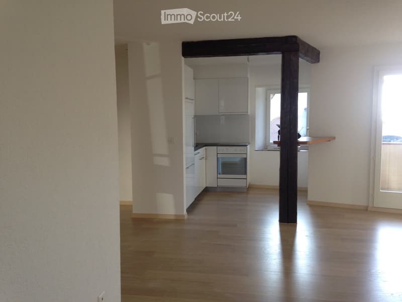 Empty room with wooden floor, white walls, kitchen with oven and refrigerator, cabinets, and a window.