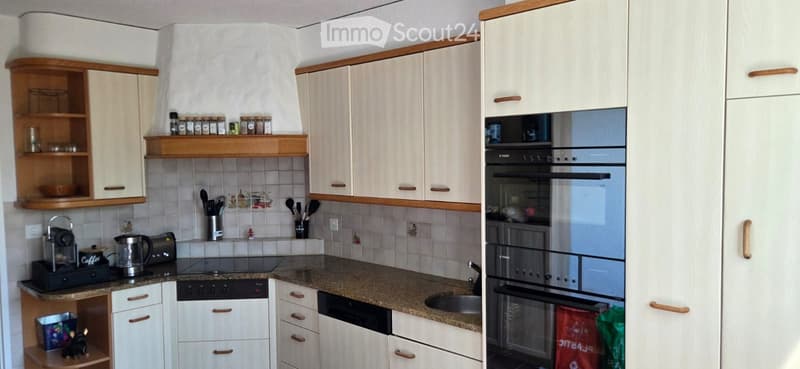 Modern kitchen with white cabinets, granite countertop, built-in oven, dishwasher, and various appliances including a coffee maker and kettle. Spice rack and utensils are visible on the walls.