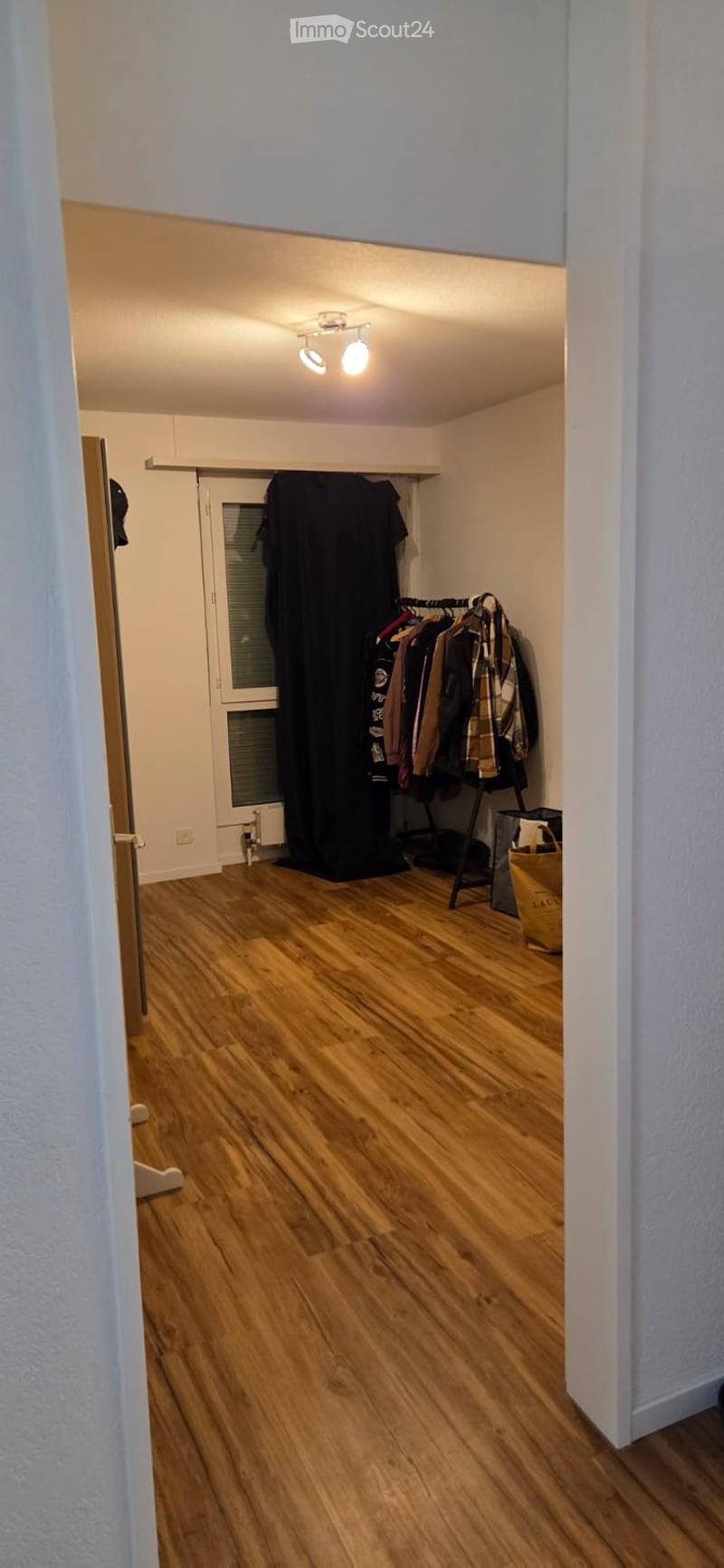Empty room with wooden flooring, white walls, and a closet with hanging clothes. The room is well-lit with ceiling lights.