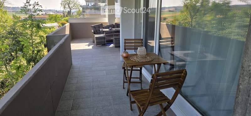 Balcony with tiled floor, wooden table and chairs, sofa and ottoman, scenic countryside view.