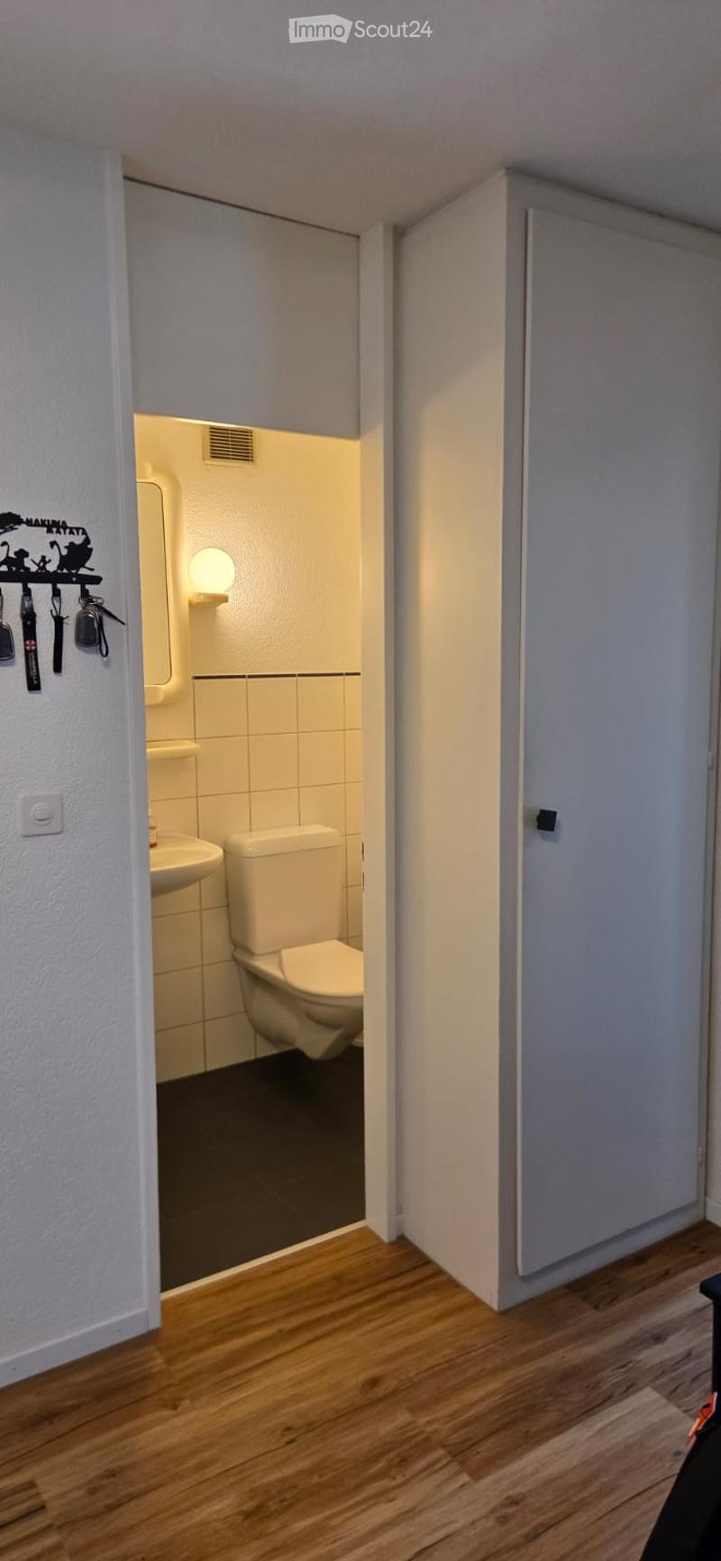 Small bathroom with white tiled walls, a toilet, sink, and a mirror. The room is illuminated by a wall-mounted light fixture.