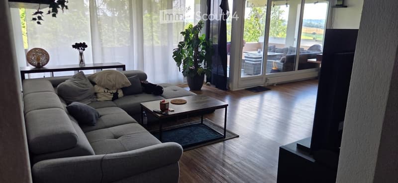 Modern living room with large windows, grey sectional sofa, coffee table, ottoman, and a view of the outdoor area.