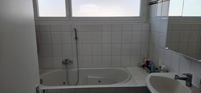Bathroom with a bathtub, sink, mirror, and tiled walls. The room has a window and a shower hose attached to the wall.