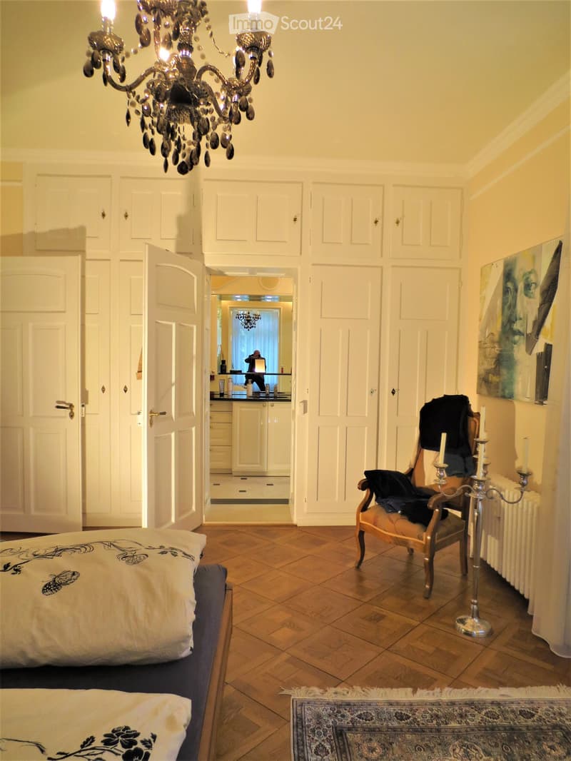 Elegant bedroom with wooden flooring, a large bed with decorative pillows, a wooden armchair with a jacket draped over it, a candelabra, a painting on the wall, a mirror reflecting a kitchen area, and a patterned rug.