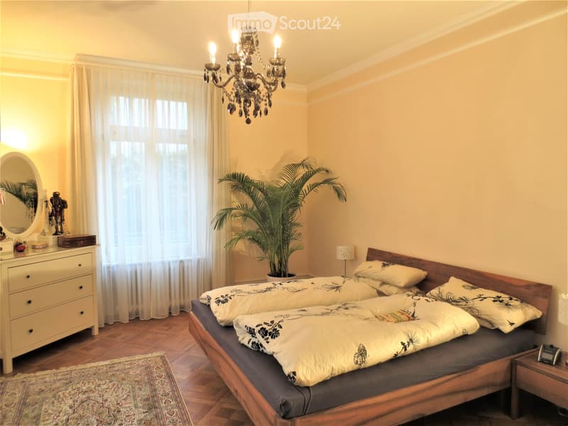 Elegant bedroom with wooden bed, patterned bedding, chandelier, dresser, mirror, and potted plant near the window.