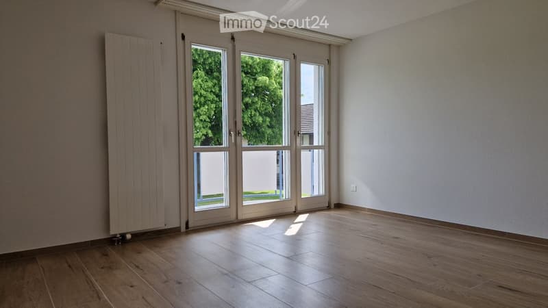 Empty room with wooden flooring, white walls, large sliding glass doors leading to an outdoor area, and a radiator on the left side.