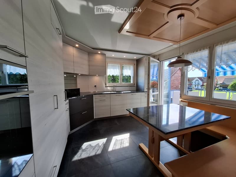 Modern kitchen with light-colored cabinets, a central island, and a dining area with a black countertop table and wooden chairs. The space features large windows and a pendant light hanging from the ceiling.