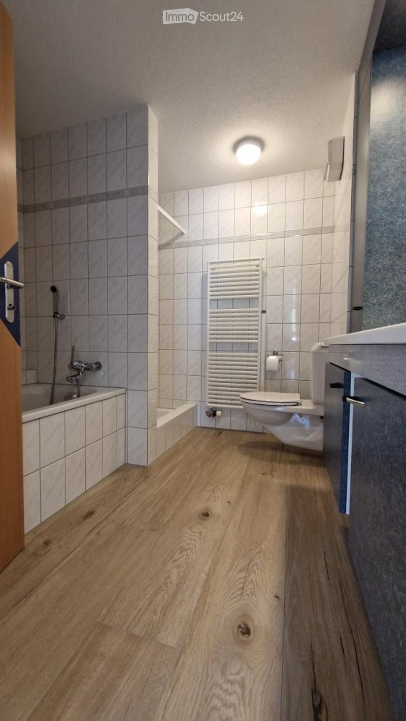 Modern bathroom with light grey tiled walls, wooden flooring, a bathtub, toilet, and a white radiator.