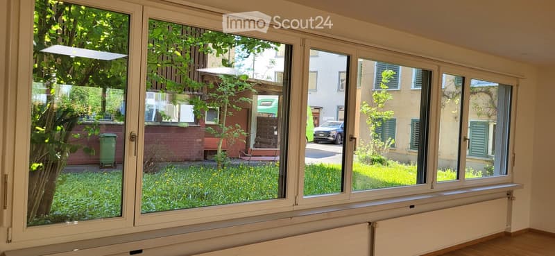 Empty room with large windows showing a view of a garden and neighboring buildings. The room has wooden flooring and a radiator below the windows.