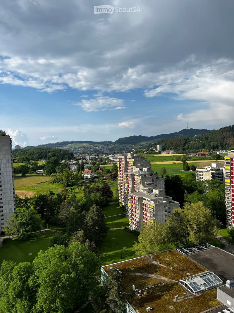 Aerial view of a residential area with multiple apartment buildings, green spaces, and a distant cityscape under a partly cloudy sky.