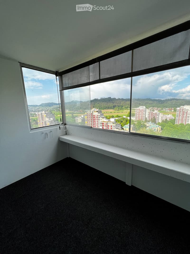 Empty room with large windows offering a view of a cityscape and surrounding landscape. The room features a white bench along the window sill and a dark carpeted floor.