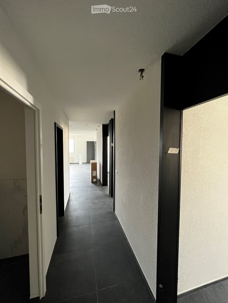 A narrow hallway with tiled flooring, white walls, and several closed doors on both sides. The hallway leads to an open area with a visible piece of furniture in the distance.