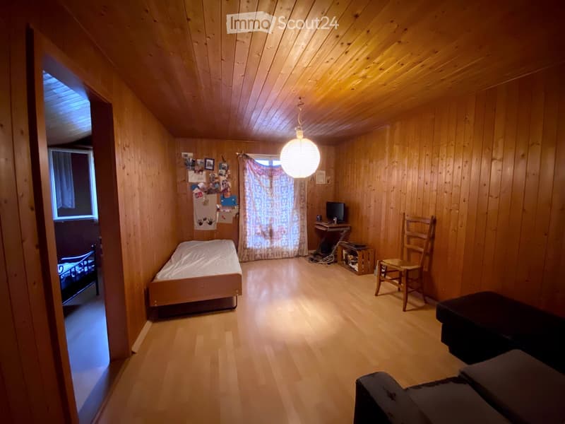 Cozy wooden bedroom with a single bed, a small table with a computer, a wooden chair, and a sofa. The room features wooden walls and ceiling, a hanging light fixture, and a window with a curtain.