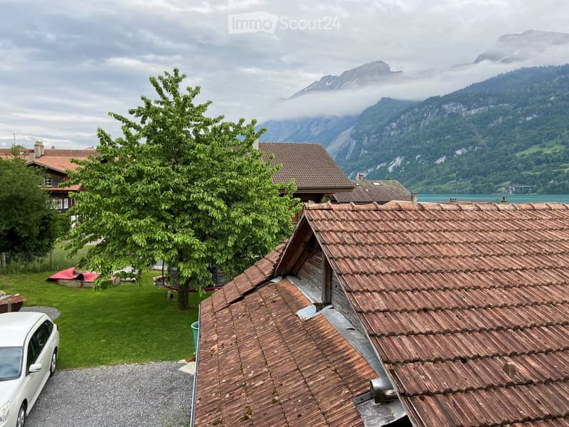 View from a rooftop showing a house with a tiled roof, a car parked nearby, a well-maintained lawn, and distant mountains partially shrouded in mist.