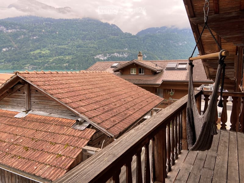 Wooden balcony with a hanging chair, overlooking a scenic lake and mountain landscape with traditional wooden houses.