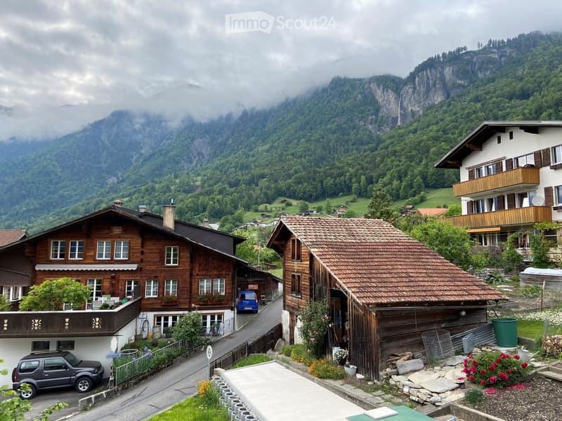 Traditional wooden houses with tiled roofs in a mountainous village setting, surrounded by lush greenery and misty mountains.