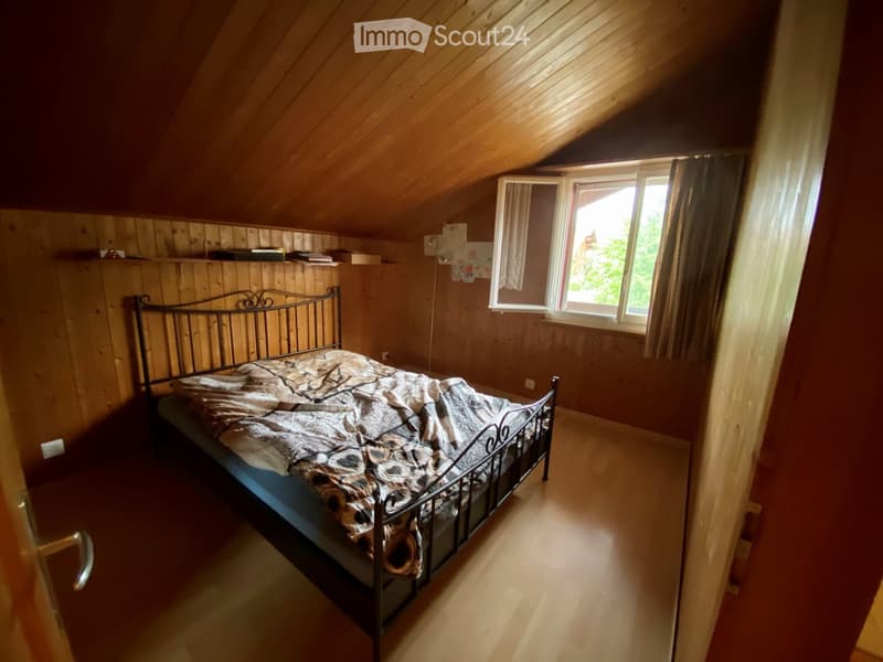 Cozy attic bedroom with wooden walls and slanted ceiling, featuring a metal-framed bed with patterned bedding and a window with curtains.