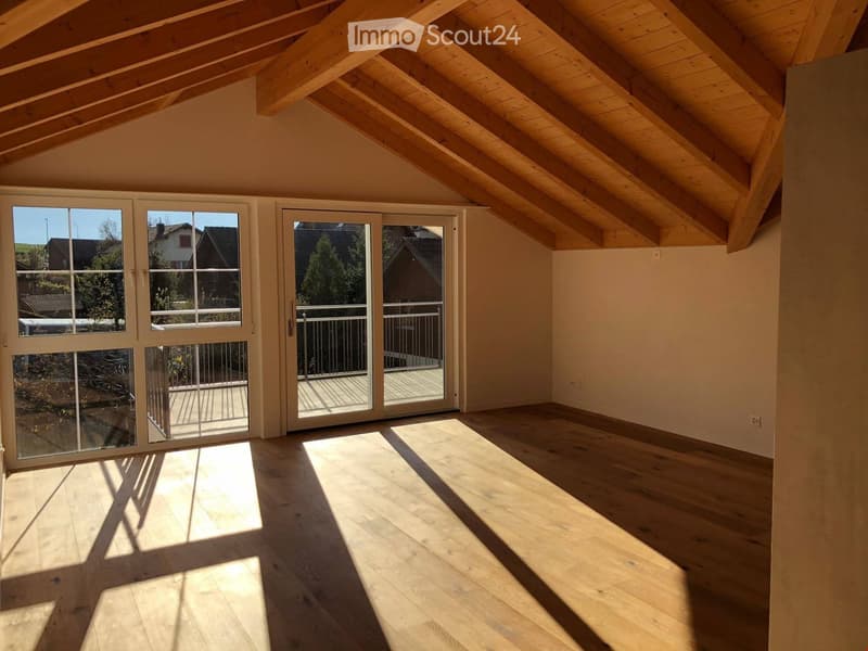 Bright, empty room with wooden flooring and exposed wooden beams on the ceiling. Large sliding glass doors lead to a balcony with a metal railing.