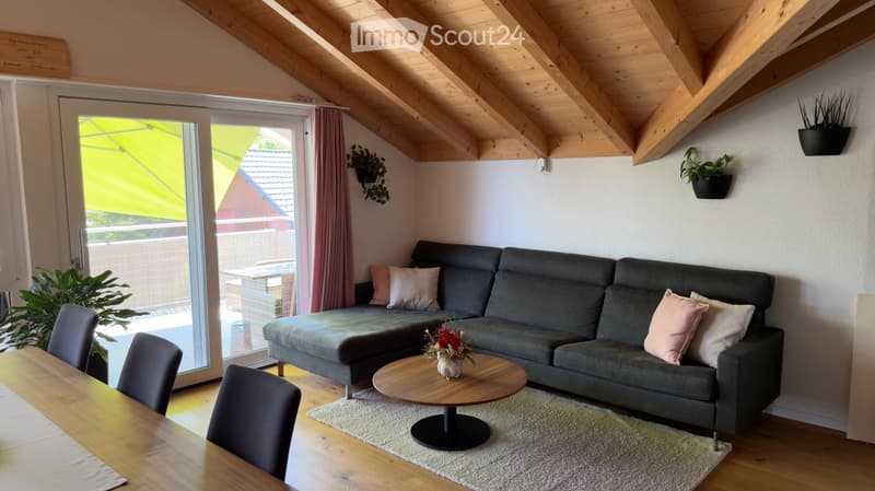 Living room with grey sectional sofa, wooden coffee table, dining table, and chairs. Exposed wooden ceiling beams and large sliding glass door leading to outdoor area.