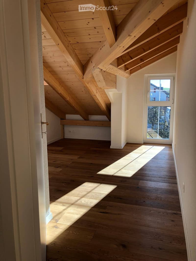 Bright, airy hallway with wooden floors and exposed wooden beams on the ceiling. Natural light streams in from a window at the end, casting shadows on the floor.