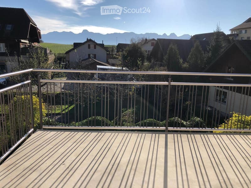 Balcony with metal railing, overlooking a residential area with houses, trees, and distant mountains.
