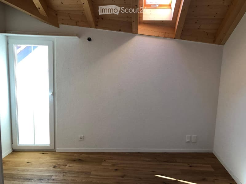 Empty room with wooden flooring, white walls, and a large window with blinds. The room has a skylight and a door leading outside.
