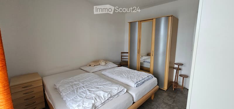 A well-lit bedroom with two single beds, a large mirrored wardrobe, a small wooden chair, a drawer unit, and a small round stool. A stuffed animal is placed on one of the beds.